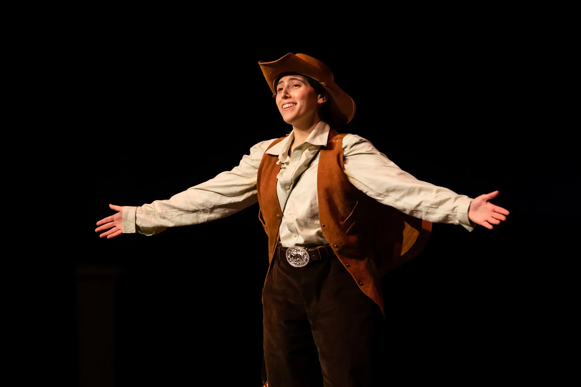 Photo of Luca Torrens wearing a cowboy hat, a tan colored long sleeved shirt and a brown vest. Their arms are outstretched and they are smiling.
