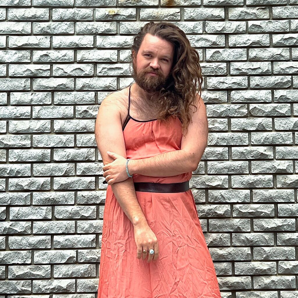 Photo of Eli Matilda Ridley. They are standing in front of a grey brick wall. They have long curly brown hair that is parted to their left side. The hair is flowing over their shoulder. The have a beard and mustache, as well as hairy chest and arms. They are wearing a pink thin strapped dress that looks a bit wrinkled. There is black trim on the dress and a black belt at the waist. Eli also has a turquoise bracelet on their left wrist and two large rings on their right hand.